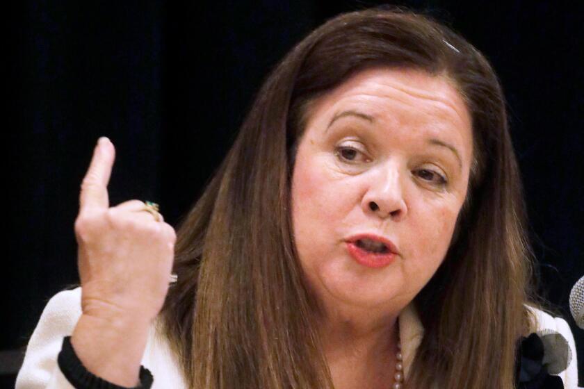 A vertical frame of a woman with medium-length brown hair in a white suit seated, gesturing with right hand while speaking