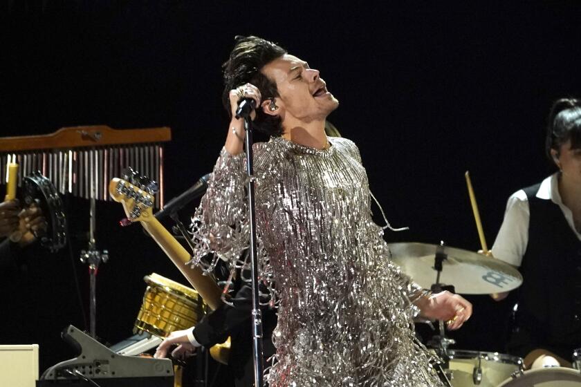 Harry Styles in a sparkly, silver fringe body suit singing on a stage and holding a microphone in his right hand