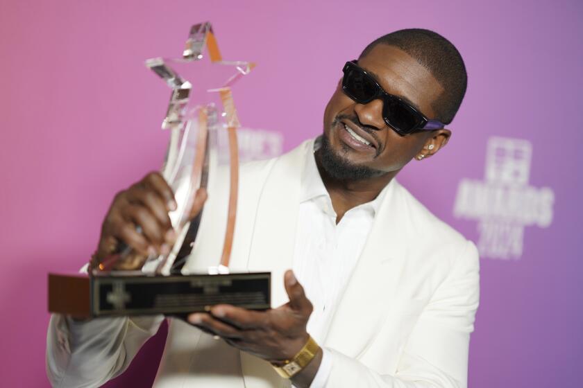 Usher in a white suit with big sunglasses smiling and holding a star-shaped glass award against a pink and purple backdrop