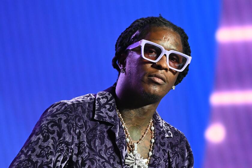 A Black man with thick, white glasses, a dark patterned shirts and chains leaning forward and looking ahead